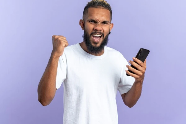 Jovem americano africano em uma camiseta branca segurando o punho cerrando o smartphone feliz e animado gritando e regozijando se com seu sucesso em pe sobre um fundo azul 141793 118821