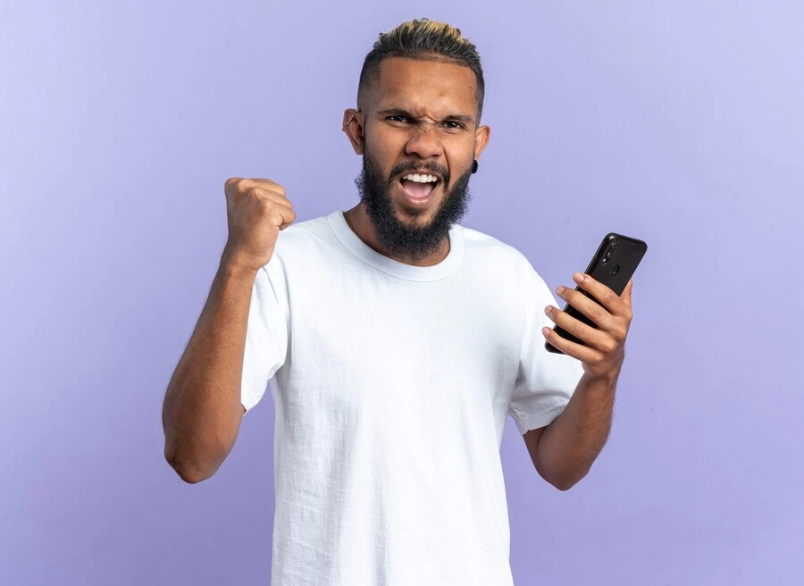 Jovem americano africano em uma camiseta branca segurando o punho cerrando o smartphone feliz e animado gritando e regozijando se com seu sucesso em pe sobre um fundo azul 141793 118821